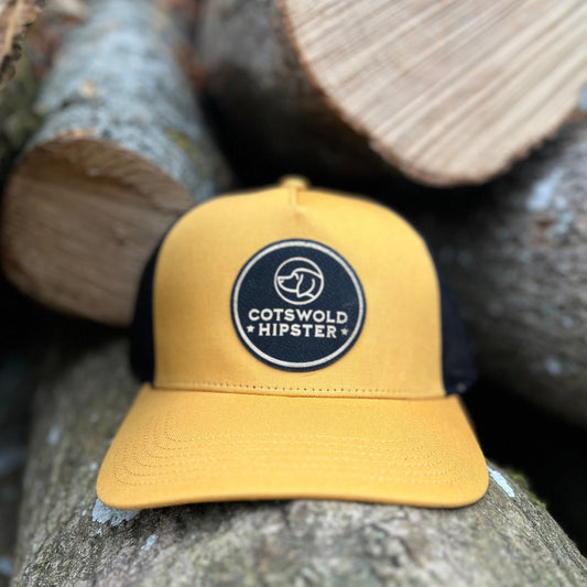 Front-facing mustard Field Cap resting on rustic logs in the Cotswolds, showing Cotswold Hipster logo and breathable mesh panels.