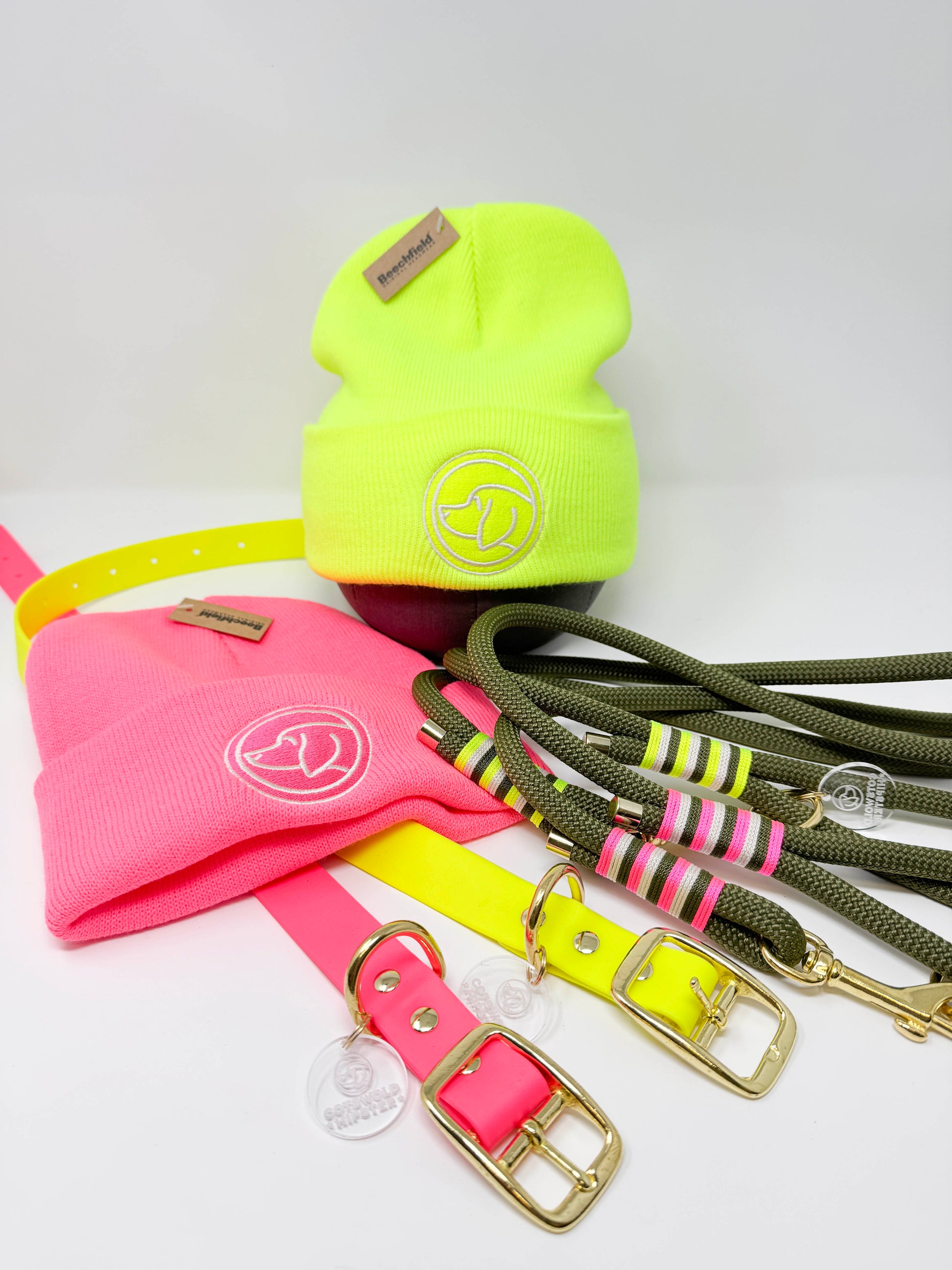 BE SEEN! collection flat lay: neon pink and neon yellow BioThane® dog collars, matching rope leads with contrast whipping, and coordinating beanie hats, all branded with dog logo, unisex and high-visibility winter dog gear.
