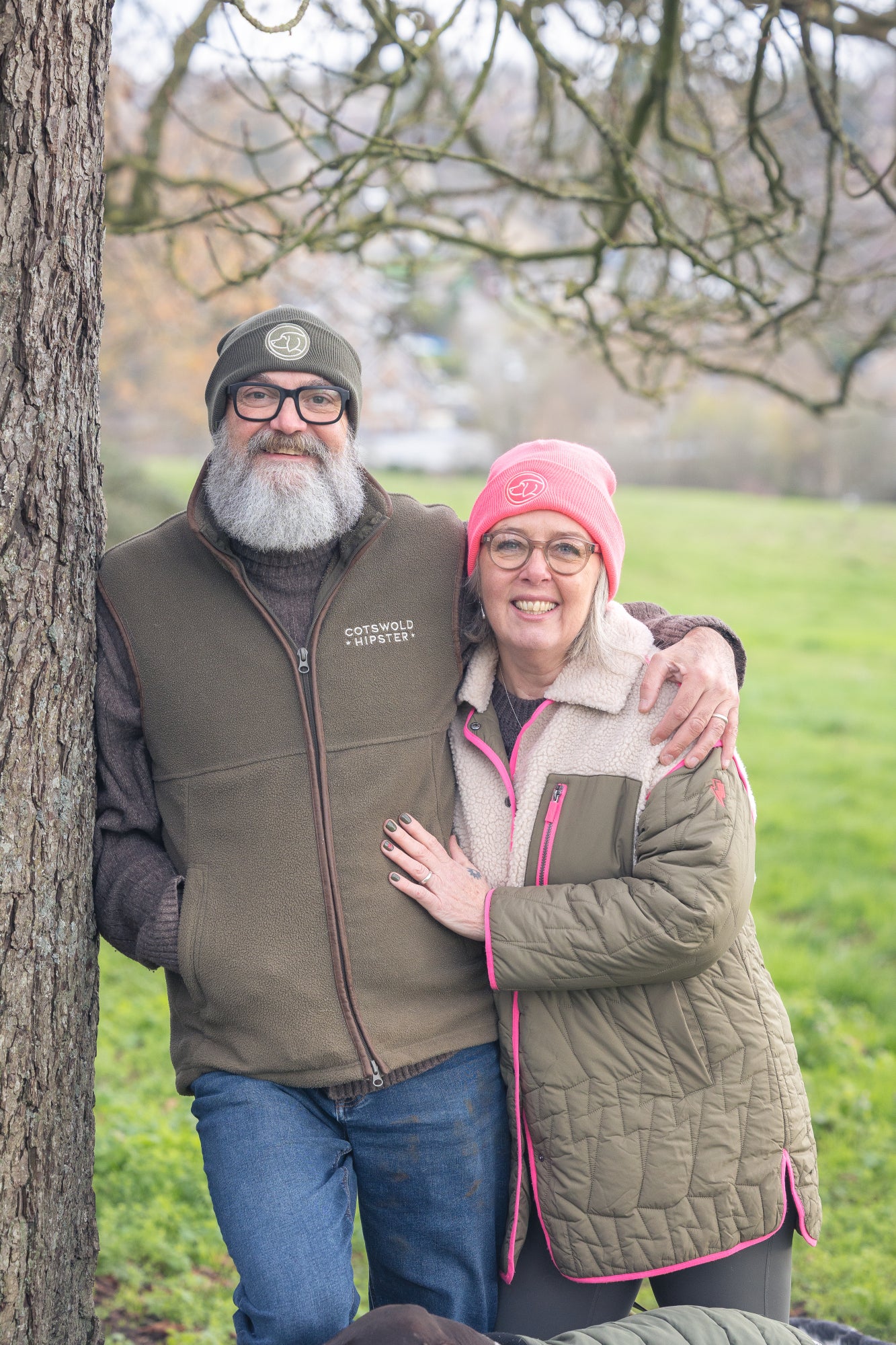 Two people wearing beanies in Cotswold  Green and Neon Pink, leaning against a tree, winter-ready coordinated style for dog owners