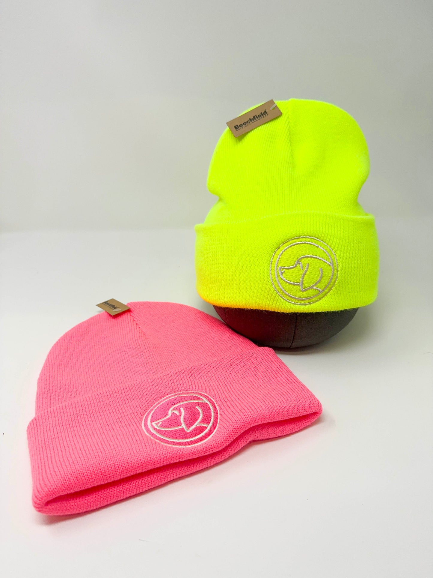 Two beanies, one neon pink and one neon yellow, with a dog head Cotswold Hipster  logo on a light gray background.