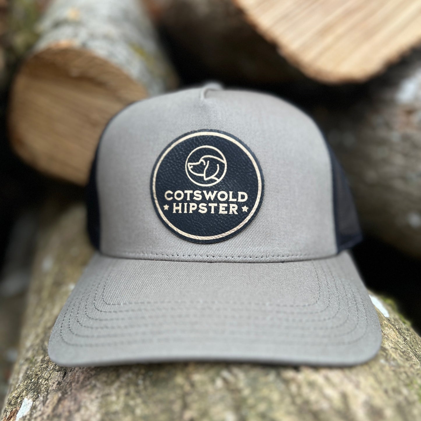 Front-facing khaki Field Cap resting on rustic Cotswold logs, showing Cotswold Hipster logo and breathable mesh panels.