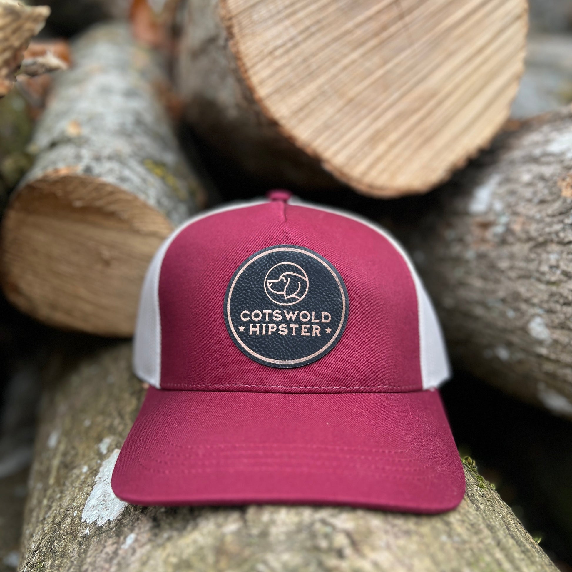 Front-facing mulberry Field Cap on rustic logs in the Cotswolds, showing Cotswold Hipster logo and breathable mesh panels.