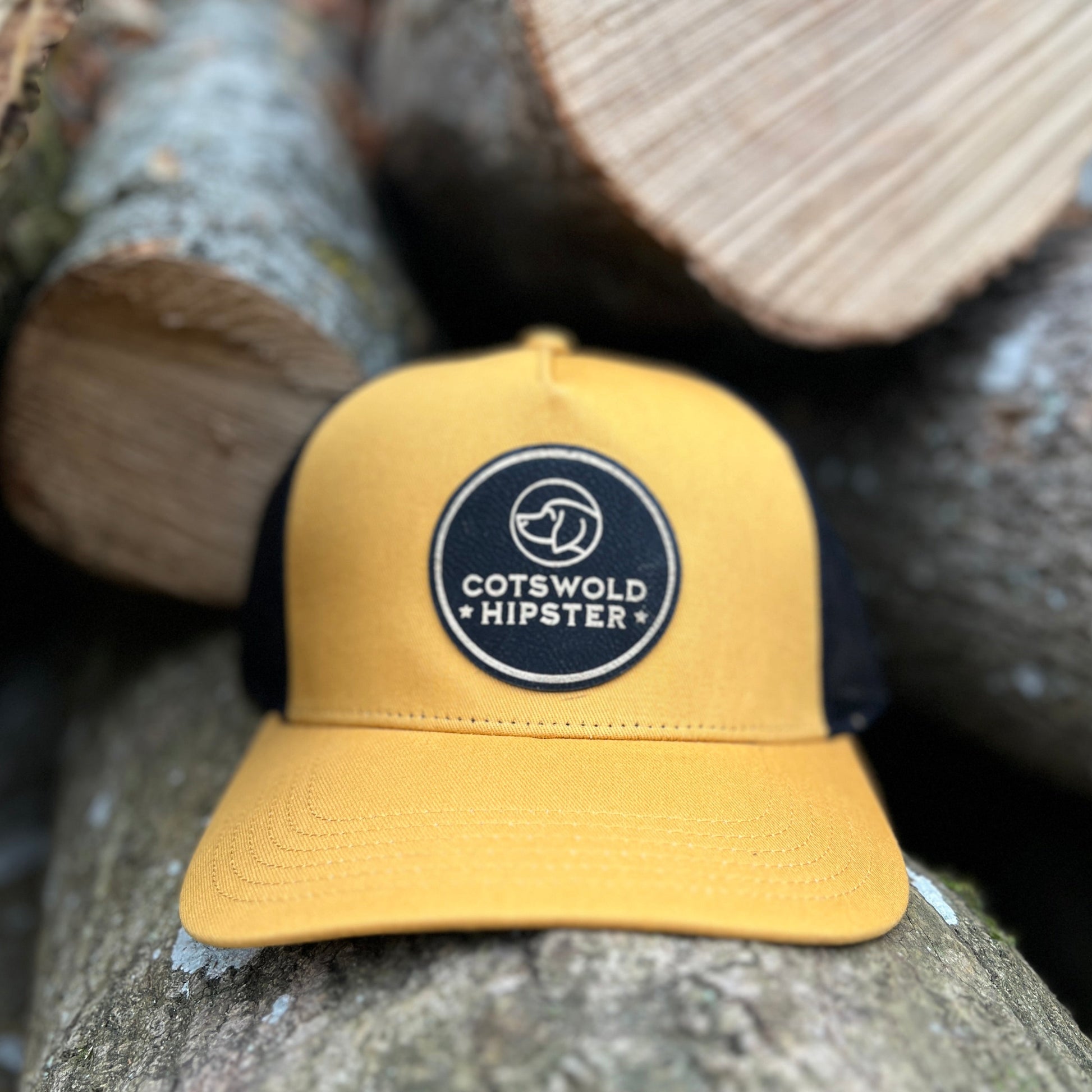 Front-facing mustard Field Cap resting on rustic logs in the Cotswolds, showing Cotswold Hipster logo and breathable mesh panels.