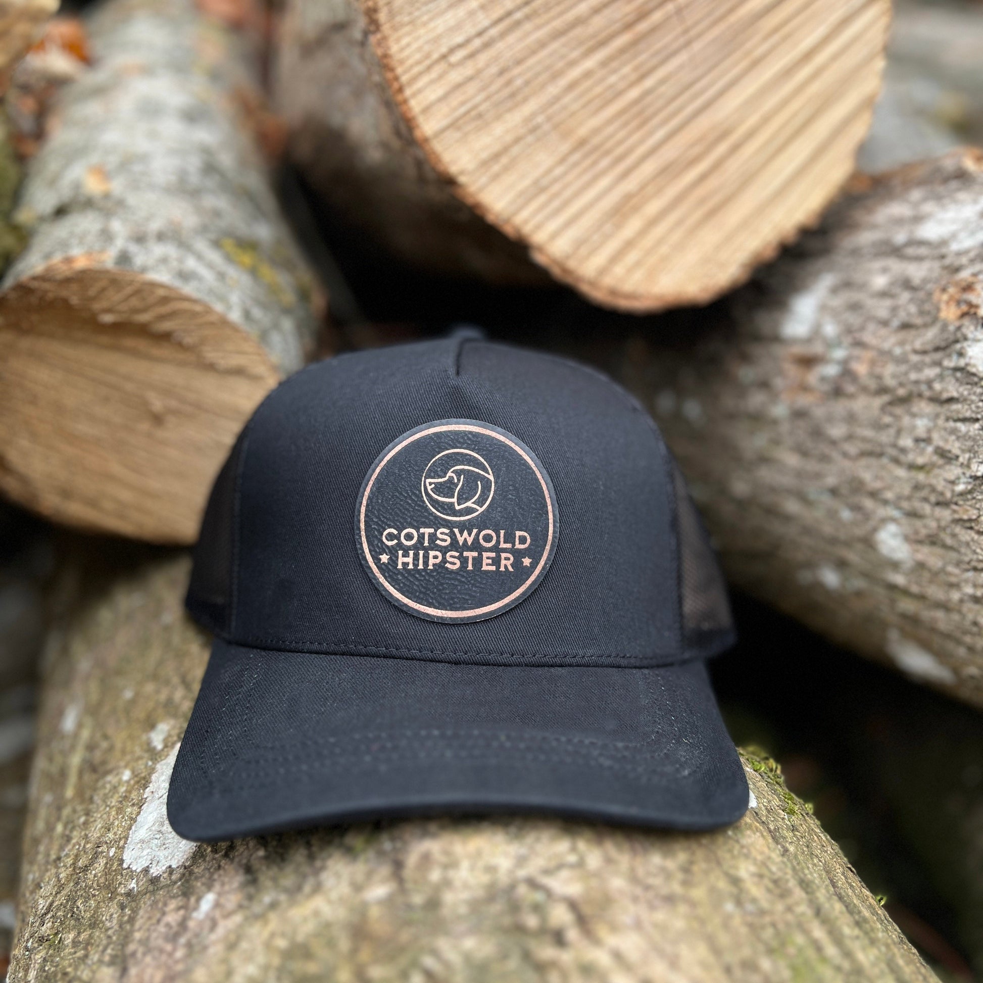 Front-facing black Field Cap resting on rustic Cotswold logs, showing Cotswold Hipster logo and breathable mesh panels.